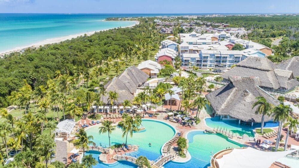 Vista aerea del resort Adult Only Memories Jibacoa con diverse piscine, palme, edifici con tetti di paglia, lettini e una lunga spiaggia di sabbia bianca accanto all'acqua turchese dell'oceano.