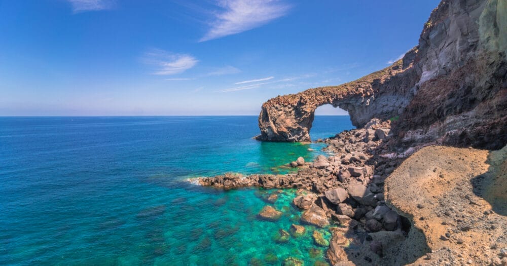 Lungo la costa frastagliata delle Isole Eolie, un arco di roccia naturale si protende su un'acqua limpida e turchese, sotto un cielo blu brillante con nuvole sottili: uno spettacolo indimenticabile durante qualsiasi tour delle Isole Eolie.